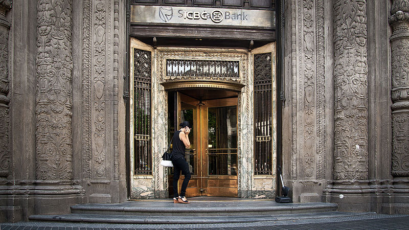 Person on phone walking into a wooden revolving door. Door is on an elaborately carved marble bank building.