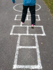 Image showing hopscotch painted on the ground, with child leaping from square to square. Child is wearing pink sneakers, balck pants and a blue jacket. We can see only the lower half of the child. There are leaves scattered around on the pavement.