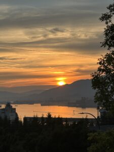 Photo taken from the point of view of Hasting Sunrise looking out over Burrard Inlet. The residences in the foreground are in silhouette against the water, which reflects the orange of the sunset sky.