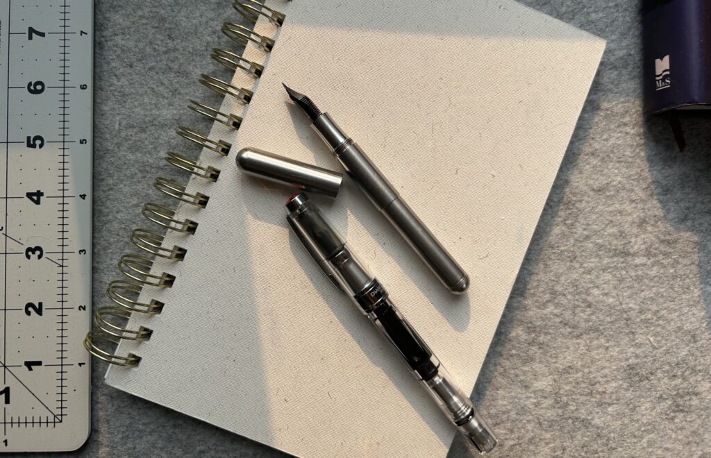 Photo of a 9 x 6 inch sketchbook with two fountain pens on it. On is a TWSBY, the other an uncapped Kaweco. The background is grey wool with a cutting mat edge showing to the left and book spines to the right.